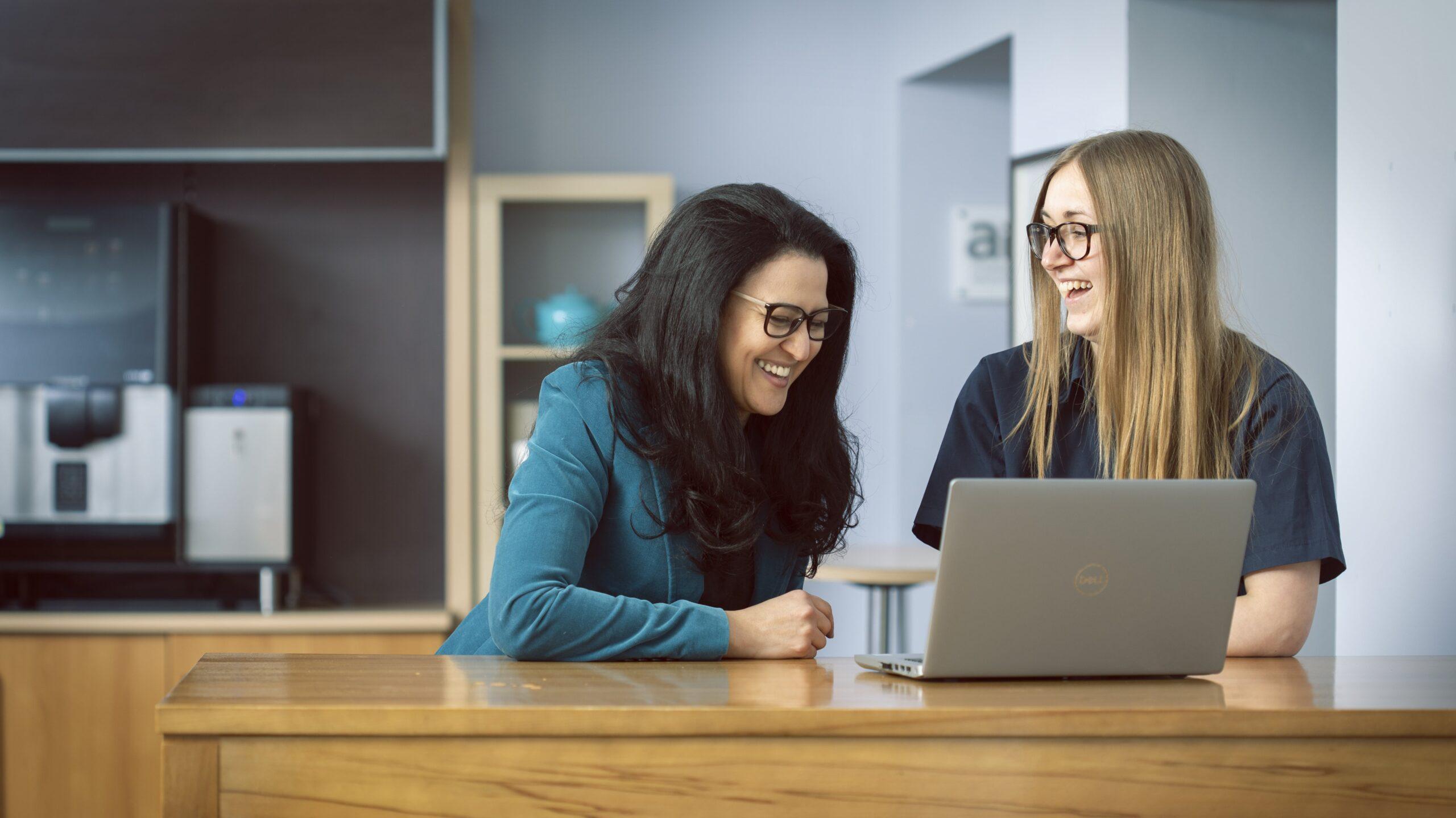 Two women with glasses smiling while watching something on a laptop together.