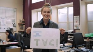 A young, smiling girl holding a handwritten poster that says "I am TVC!"